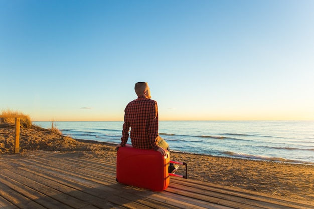 Holiday, travel and summer vacation concept -a man sitting on red suitcase and watching the sunset.