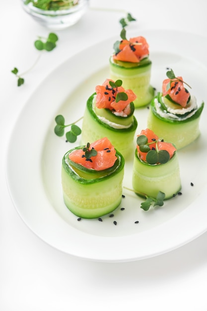 Holiday vegetable appetizers. cucumbers rolls with soft cheese, pieces of salted salmon, microgreens and black sesame served on a white plate. selective focus.