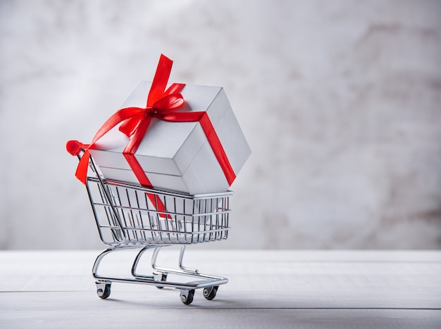 Holiday white box with red ribbon in shopping cart on gray background. Front view and copy space