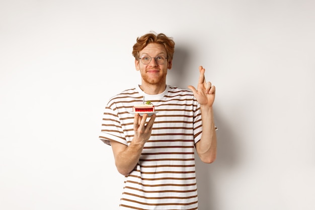 休日とお祝いのコンセプト。キャンドルでバースデーケーキを保持し、幸運と願い事を作るために指を交差させる、白い背景のメガネで陽気な赤毛の男。