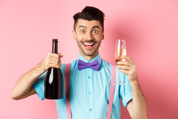 休日とお祝いのコンセプト。楽しんで、シャンパンとガラスのボトルを見せて、イベントで乾杯を作って、笑顔でカメラを見て、ピンクの背景で幸せな若い男。