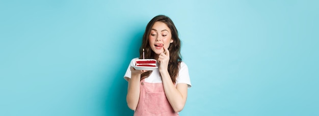 Photo holidays and celebration cute birthday girl looking at tasty cake with temptation to bite it standin