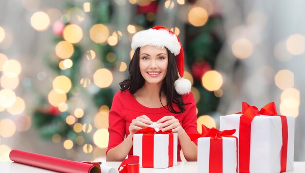 휴일, 축하, 장식 및 사람 개념 - 크리스마스 트리 조명 배경 위에 종이 포장 선물 상자를 장식하는 산타 도우미 모자를 쓴 웃는 여자