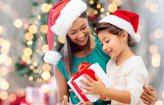 Concetto di vacanze, celebrazione, famiglia e persone - madre felice e bambina in cappelli di babbo natale con confezione regalo sopra il soggiorno e lo sfondo dell'albero di natale