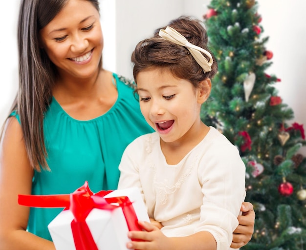 휴일, 축하, 가족 및 사람 개념 - 거실과 크리스마스 트리 배경 위에 선물 상자가 있는 행복한 어머니와 어린 소녀