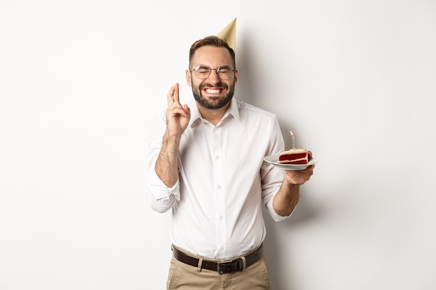 休日とお祝い。誕生日ケーキ、クロスフィンガー、興奮した笑顔、b-dayパーティー、白い背景で願い事をする幸せな男。