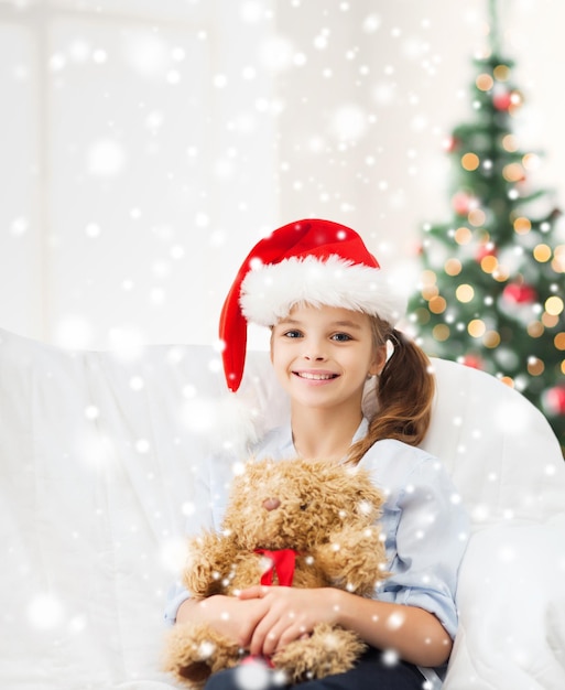 holidays, childhood and people concept - smiling girl in santa helper hat with teddy bear toy over living room with christmas tree and snow background