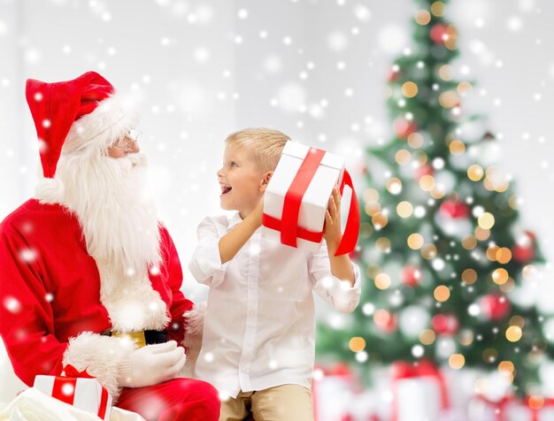 holidays, childhood and people concept - smiling little boy, santa claus with gifts over living room, christmas tree and snow background