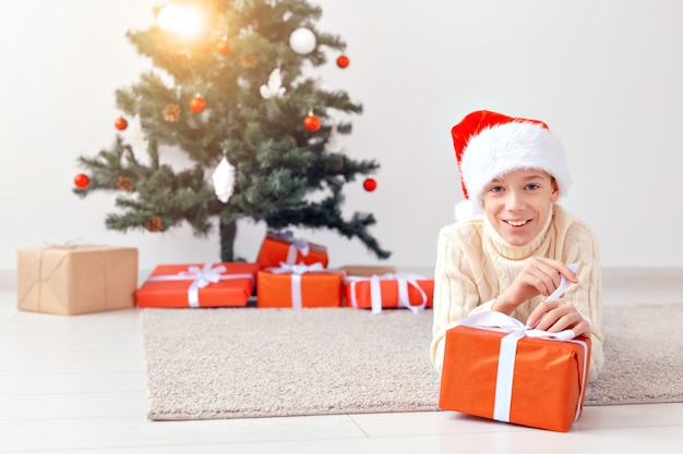 Holidays, christmas, childhood and people concept - smiling happy teen boy in santa hat opens gift box over christmas tree background.