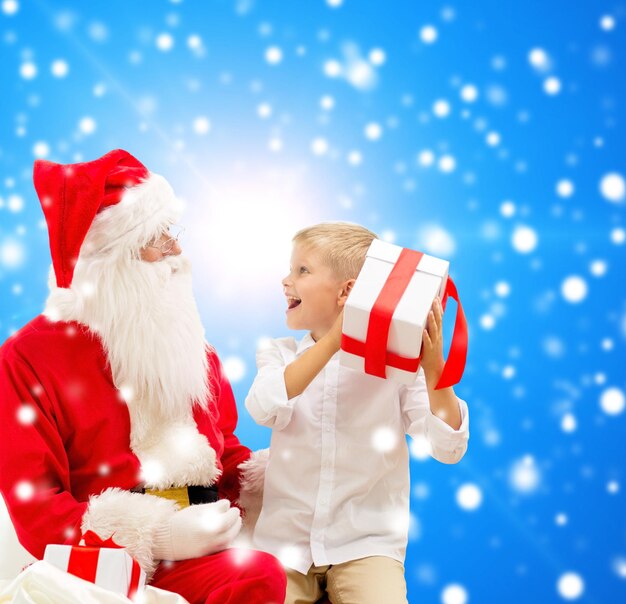 Photo holidays, christmas, childhood and people concept - smiling little boy with santa claus and gifts over blue snowy background