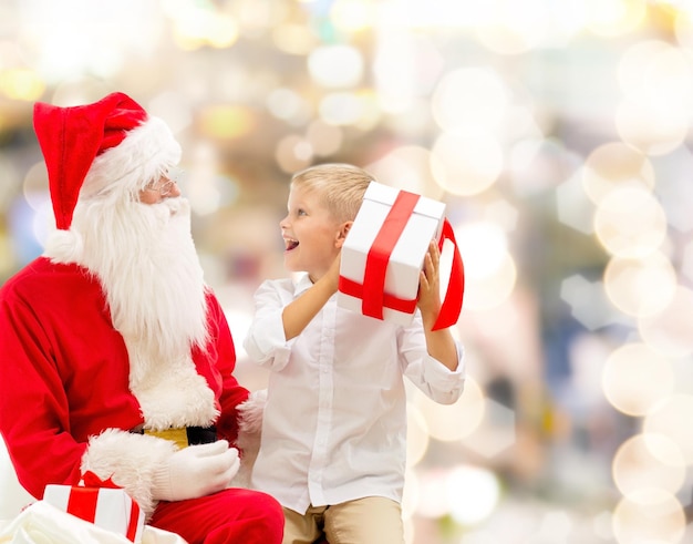 holidays, christmas, childhood and people concept - smiling little boy with santa claus and gifts over lights background