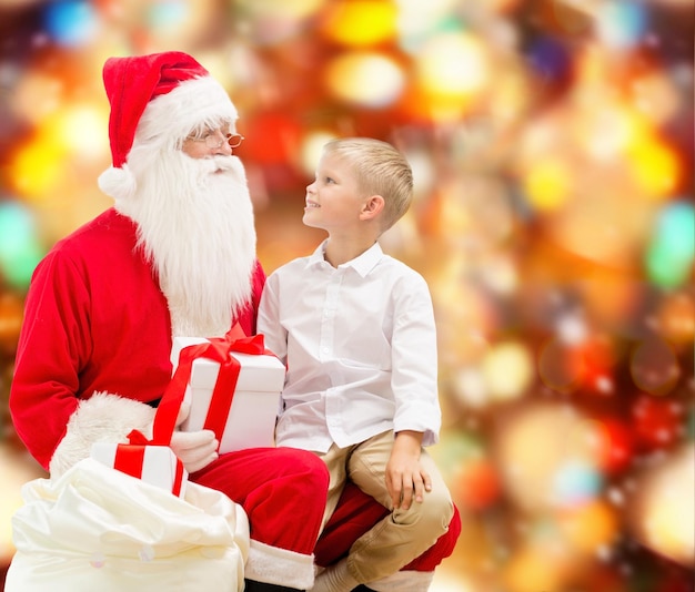 праздники, рождество, детство и концепция людей - улыбающийся маленький мальчик с дедом морозом и подарками на фоне красных огней