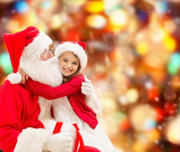 holidays, christmas, childhood and people concept - smiling little girl hugging with santa claus over red lights background