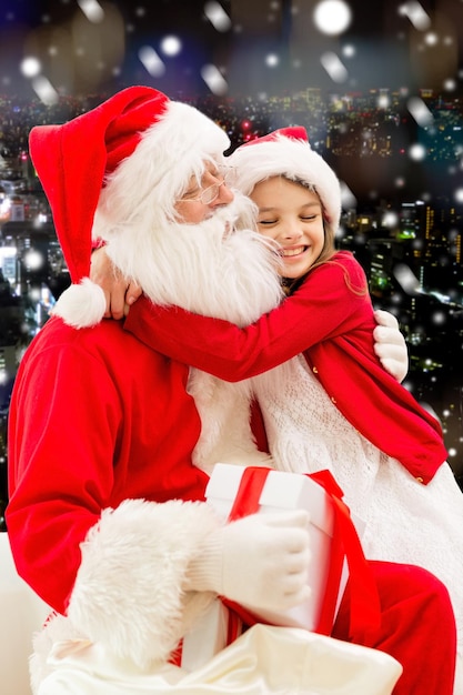 Photo holidays, christmas, childhood and people concept - smiling little girl hugging with santa claus over snowy city background