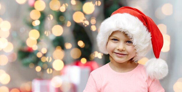 holidays, christmas, childhood and people concept - smiling little girl in santa hat sitting on couch over lights background