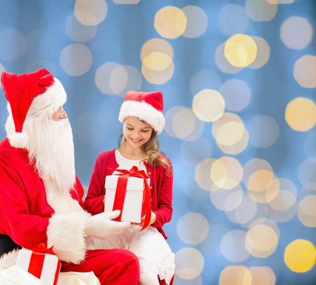 holidays, christmas, childhood and people concept - smiling little girl with santa claus and gifts over blue lights background