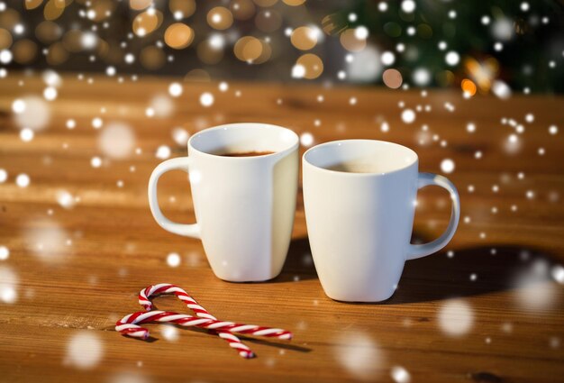 holidays, christmas, winter, food and drinks concept - close up of candy canes and cups with hot chocolate or cocoa drinks on wooden table over christmas tree lights