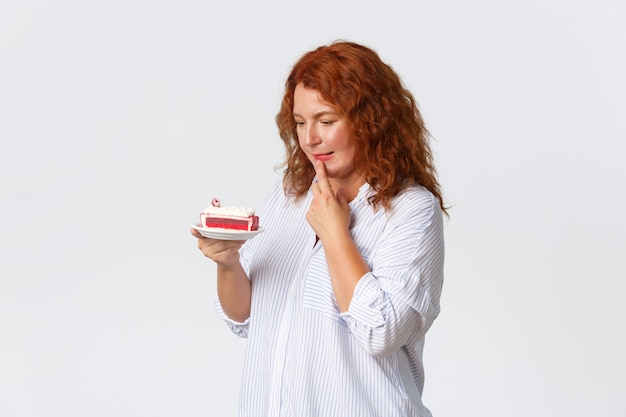 Фото Праздники, эмоции и концепция образа жизни. нерешительная милая женщина средних лет с рыжими волосами принимает решение, хочет съесть торт, но беспокоится о калориях и внимательно смотрит на десерт.