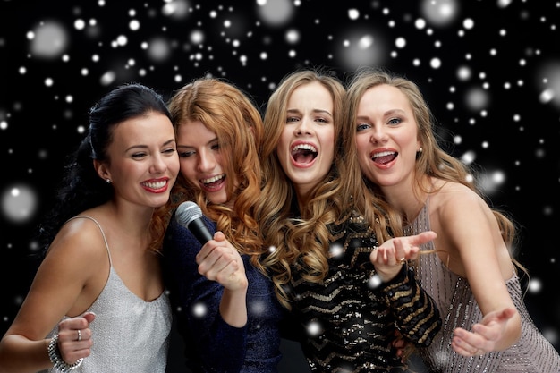 holidays, friends, bachelorette party, nightlife and people concept - three women in evening dresses with microphone singing karaoke over black background