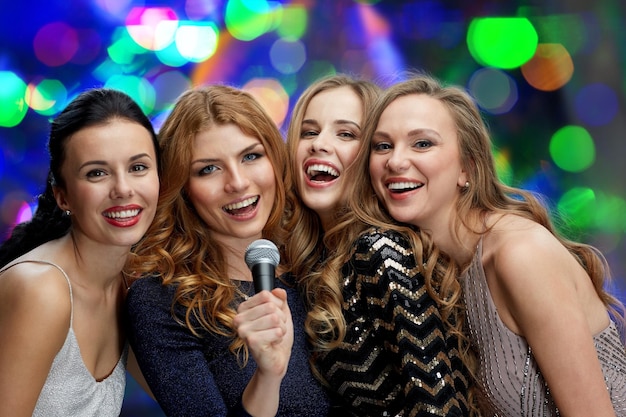 Foto vacanze, amici, addio al nubilato, vita notturna e concetto di persone - tre donne in abiti da sera con microfono che cantano karaoke su sfondo di luci
