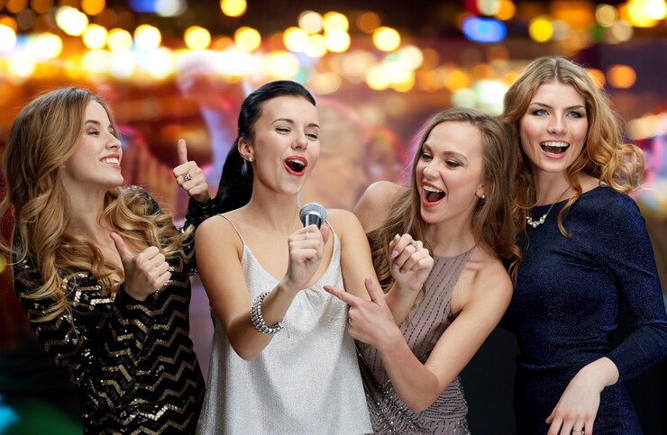 Photo holidays, friends, bachelorette party, nightlife and people concept - three women in evening dresses with microphone singing karaoke over night club disco lights background