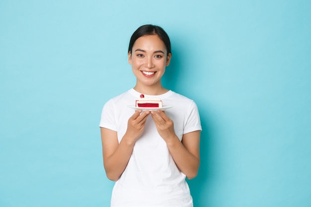 Holidays, lifestyle and celebration concept. Smiling beautiful slim asian girl in white t-shirt, showing piece of cake and looking upbeat, eating delicious dessert, standing light blue wall.