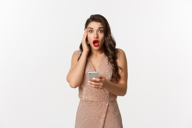 Праздники, концепция покупок в Интернете. Удивленная женщина, использующая мобильный телефон и выглядящая изумленной, в вечернем платье, стоя на белом фоне.