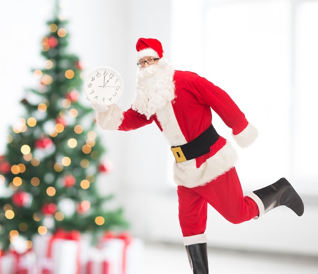 holidays and people concept - man in costume of santa claus running with clock showing twelve over living room and christmas tree background