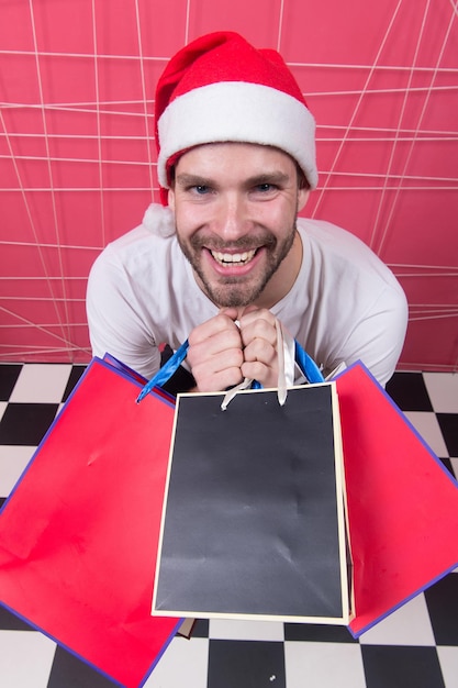 Holidays preparation and celebration. Man in xmas hat smile with colorful bags. Christmas and new year presents. Winter sale, shopping concept. Santa shopper with paperbags on pink background.