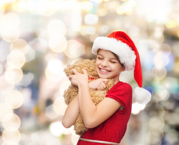 holidays, presents, christmas, childhood and people concept - smiling girl in santa helper hat with teddy bear over lights background