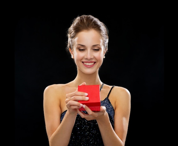 holidays, presents, luxury and happiness concept - smiling woman in dress holding red gift box over black background