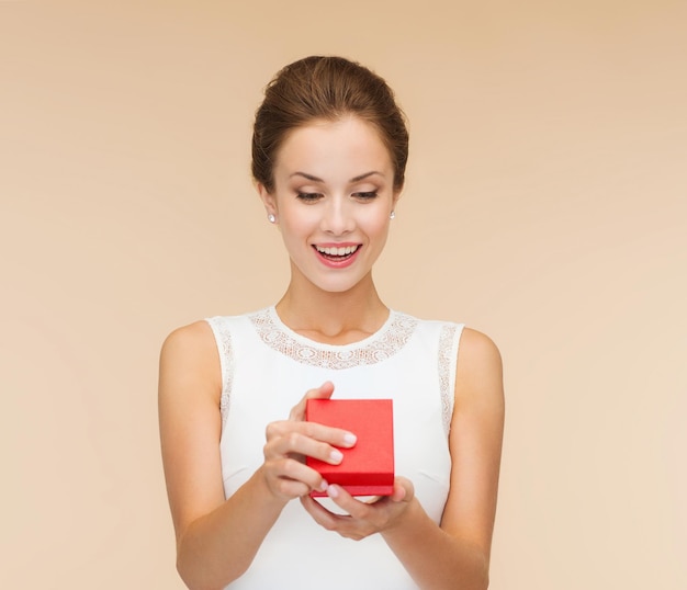 праздники, подарки, свадьба и концепция счастья - улыбающаяся женщина в белом платье с красной подарочной коробкой на бежевом фоне