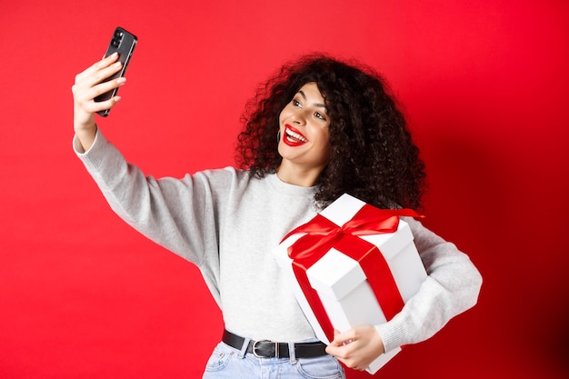 Vacanze e concetto di tecnologia. donna felice che prende selfie con il suo regalo, tenendo presente e smartphone, in piedi su sfondo rosso