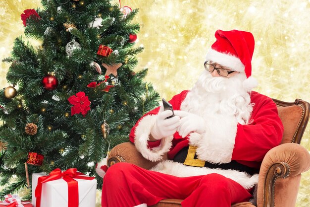 holidays, technology and people concept - man in costume of santa claus with smartphone, presents and christmas tree over yellow lights background