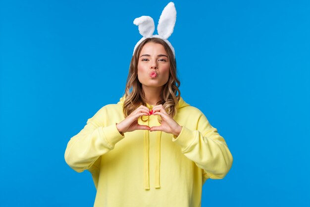 Holidays, traditions and celebration concept Funny and cute silly blond girl in yellow hoodie and rabbit ears showing heart gesture, make mwah kiss expression, spread love and positivity