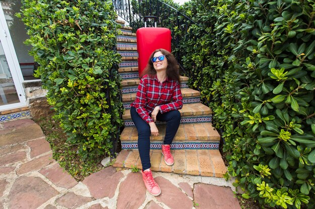 Holidays, travel, people concept. young woman in sunglasses sitting on stairs with suitcases and smiling.