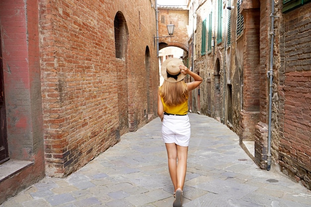 Holidays in Tuscany Back view of beautiful stylish tourist girl walking between narrow alleys of Siena historic town of Tuscany Italy