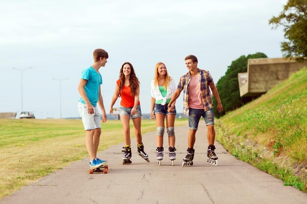 Photo holidays, vacation, love and friendship concept - group of smiling teenagers with roller skates and skateboard riding outdoors