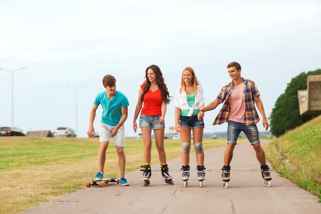 концепция праздников, каникул, любви и дружбы - группа улыбающихся подростков с роликовыми коньками и скейтбордом, катающихся на открытом воздухе