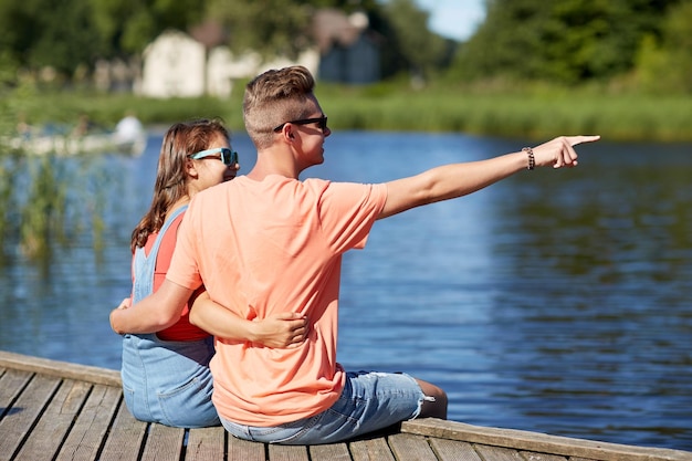 holidays, vacation, love and people concept - happy smiling teenage couple hugging and pointing finger to something on river berth at summer