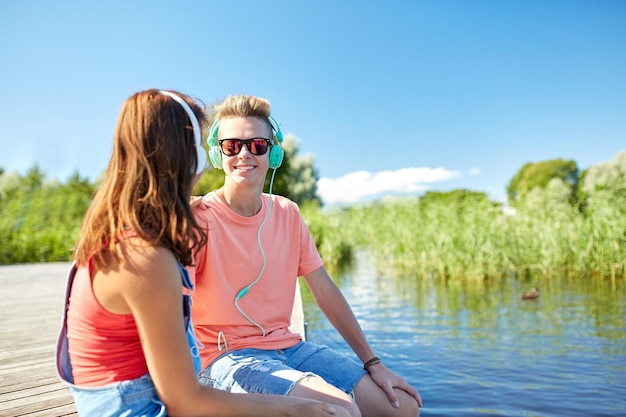 holidays, vacation, love and people concept - happy teenage couple with earphones sitting on river berth and listening to music at summer