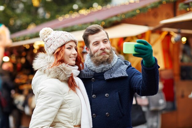 holidays, winter, christmas, technology and people concept - happy couple of tourists in warm clothes taking selfie with smartphone in old town