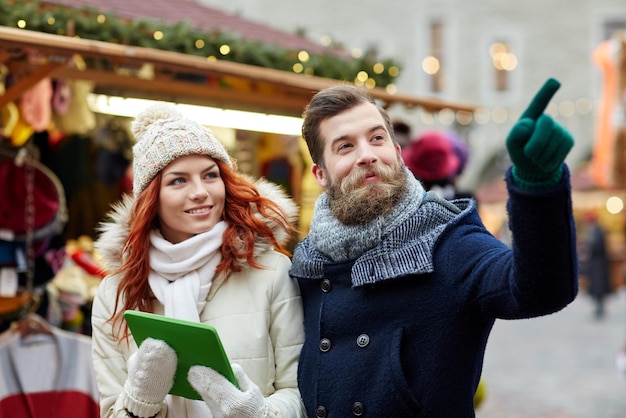 holidays, winter, christmas, technology and people concept - happy couple of tourists in warm clothes with tablet pc computer pointing finger in old town