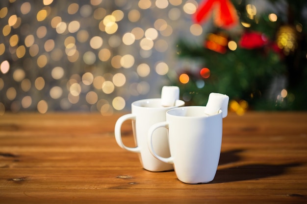 holidays, winter, food and drinks concept - close up of cups with hot chocolate or cocoa drinks and marshmallow on wooden table over lights