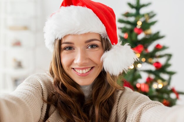 휴일, 겨울, 그리고 사람들의 개념 - 산타 모자를 쓴 행복한 젊은 여성이 집에서 크리스마스 트리 위로 셀카를 찍습니다.