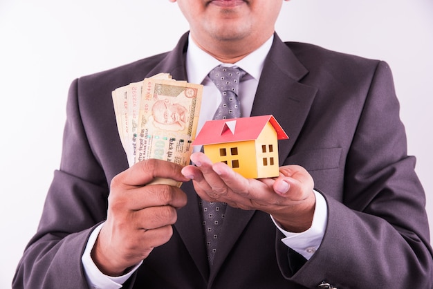 Home and car buying concept, indian man holding indian currency in both hands and model house and toy car over it, closeup and selective focus