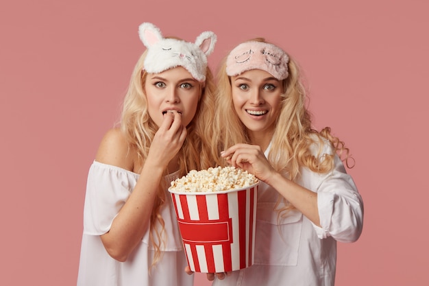 Home cinema online. Young women in pajamas and sleep masks watching a movie at home on isolation in front of a television with popcorn