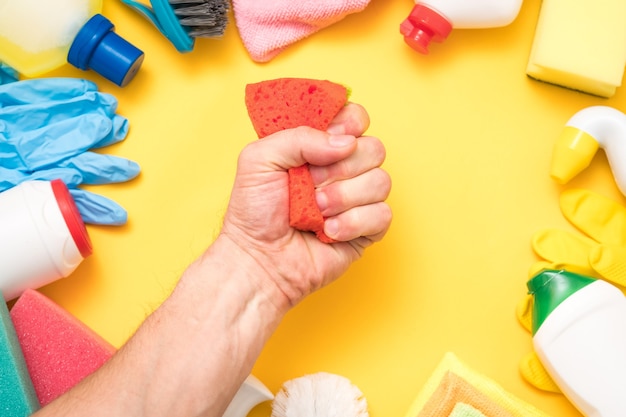 Home cleaning concept. Man hand squeezing sponge. Assorted supplies frame on yellow.