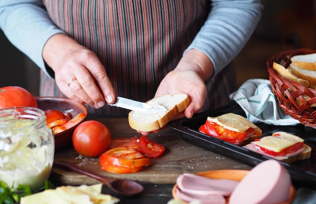 Photo home cooking elderly woman preparing hot sandwiches with cheese tomato sausage on toast