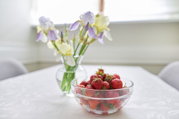 ホームダイニングルームのインテリア、白いテーブルクロスのテーブル、花瓶に菖蒲の花の春夏の花束、熟したイチゴのボウル